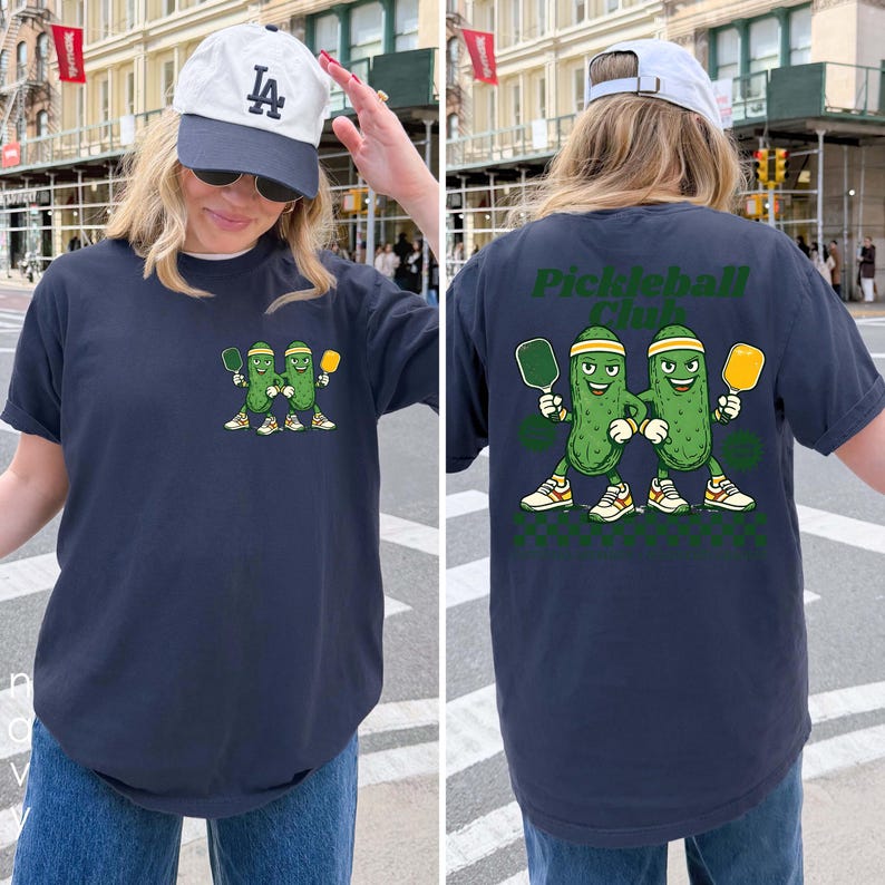 May include: A navy blue t-shirt featuring a small graphic on the front left chest and a large graphic on the back. Both graphics show two cartoon pickles wearing headbands and sneakers, holding pickleball paddles. The back includes the text Pickleball Club.