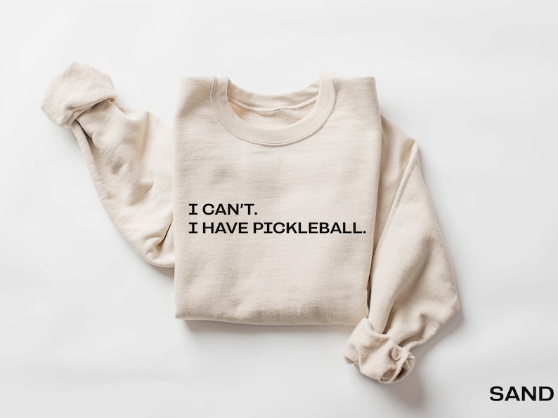 May include: A beige sweatshirt with the text 'I CAN'T. I HAVE PICKLEBALL.' printed in black on the front.