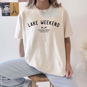 May include: An ivory short-sleeve t-shirt featuring the black text LAKE WEEKEND printed in a curved arch above a graphic of two crossed oars over stylized water waves. The shirt has a crew neckline and is paired with light blue denim jeans.