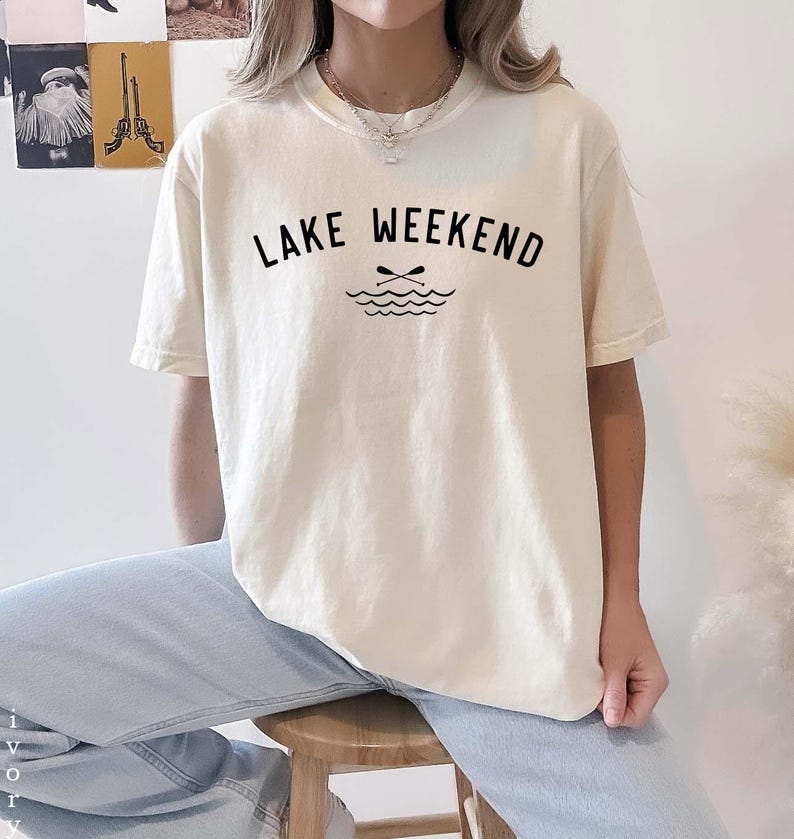 May include: An ivory short-sleeve t-shirt featuring the black text LAKE WEEKEND printed in a curved arch above a graphic of two crossed oars over stylized water waves. The shirt has a crew neckline and is paired with light blue denim jeans.