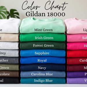 May include: A Gildan 18000 color chart displaying three vertical stacks of folded crewneck sweatshirts in various colors. The text reads Color Chart Gildan 18000, with labels for each color including White, Sand, Ash, Sport Grey, Dark Heather, Military Green, Dark Chocolate, Charcoal, Mint Green, Irish Green, Forest Green, Sapphire, Royal, Navy, Carolina Blue, Indigo Blue, Light Pink, Safety Pink, Heliconia, Red, Cardinal Red, Maroon, Purple, and Violet.