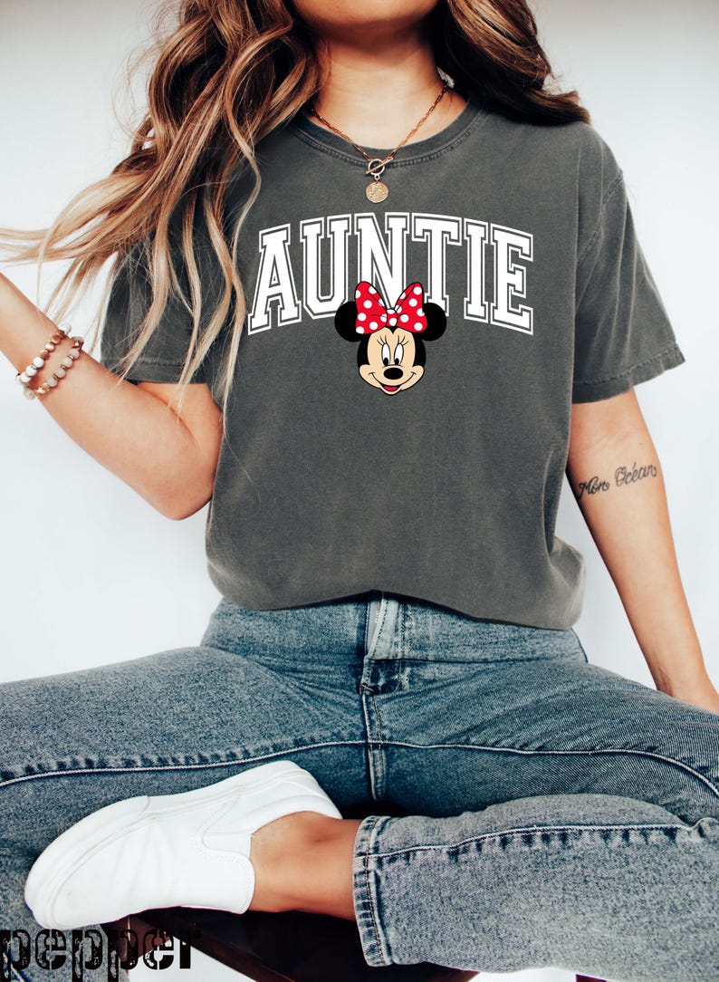 May include: Dark gray t-shirt with the word "AUNTIE" in white block letters and a Minnie Mouse graphic. The Minnie Mouse graphic features a red bow with white polka dots. The shirt is paired with blue jeans and white sneakers.