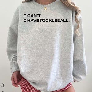 Pode incluir: Um moletom cinza claro com o texto preto "I CAN'T. I HAVE PICKLEBALL." A pessoa est&aacute; usando shorts xadrez vermelho e branco. Um colar de ouro &eacute; vis&iacute;vel.