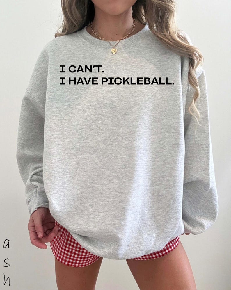 Pode incluir: Um moletom cinza claro com o texto preto "I CAN'T. I HAVE PICKLEBALL." A pessoa est&aacute; usando shorts xadrez vermelho e branco. Um colar de ouro &eacute; vis&iacute;vel.