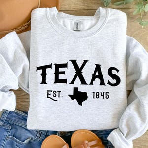 K&ouml;nnte beinhalten: Ein hellgraues Sweatshirt mit Rundhalsausschnitt, schwarzem Schriftzug TEXAS, EST. und 1845 sowie einer schwarzen Silhouette des Bundesstaates Texas. Das Kleidungsst&uuml;ck hat lange &Auml;rmel, gerippte B&uuml;ndchen am Kragen, &Auml;rmeln und Saum sowie ein Gildan-Etikett.