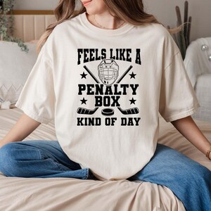 A woman wearing a white t-shirt with a humorous text printed on it, sitting on a bed.