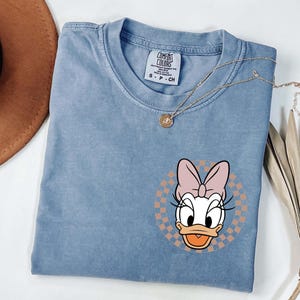 Op de afbeelding: Een lichtblauw T-shirt met een Daisy Duck-afbeelding. De afbeelding is een cartoonafbeelding van Daisy Duck met een roze strik, tegen een geruite achtergrond. Het shirt heeft een ronde hals en korte mouwen.