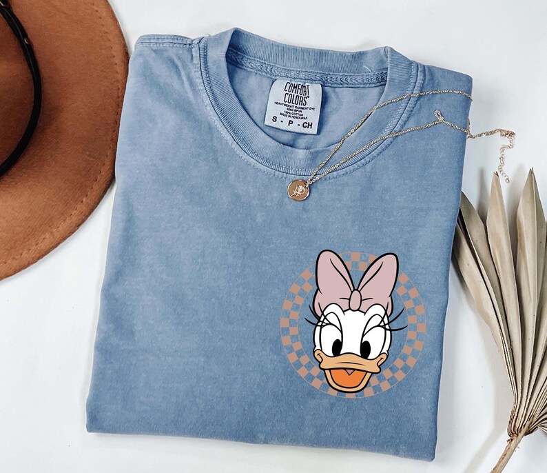 Op de afbeelding: Een lichtblauw T-shirt met een Daisy Duck-afbeelding. De afbeelding is een cartoonafbeelding van Daisy Duck met een roze strik, tegen een geruite achtergrond. Het shirt heeft een ronde hals en korte mouwen.