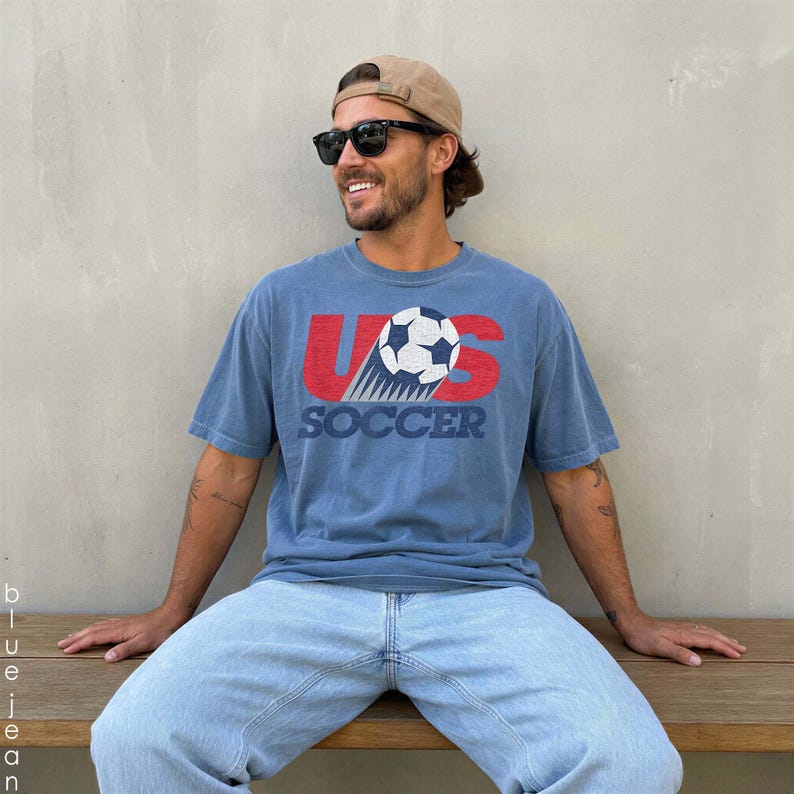 Pu&ograve; includere: Una t-shirt blu a maniche corte con una stampa grafica che presenta le lettere rosse US, un pallone da calcio bianco e nero e la parola SOCCER in blu. &Egrave; abbinata a jeans azzurri, un berretto da baseball beige e occhiali da sole neri.