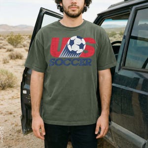 Pu&ograve; includere: Una t-shirt a maniche corte color verde oliva con una stampa grafica che include il testo US in rosso, un'illustrazione di un pallone da calcio e la parola SOCCER in blu. Il design presenta un aspetto usurato e testurizzato sul davanti.