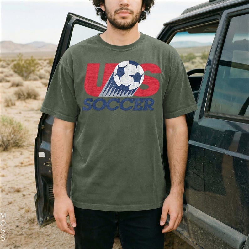 Pu&ograve; includere: Una t-shirt a maniche corte color verde oliva con una stampa grafica che include il testo US in rosso, un'illustrazione di un pallone da calcio e la parola SOCCER in blu. Il design presenta un aspetto usurato e testurizzato sul davanti.