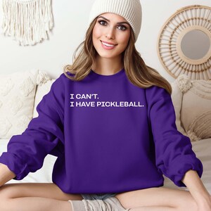 Pode incluir: Um moletom roxo escuro com o texto branco "I CAN'T. I HAVE PICKLEBALL.". A parte superior de manga comprida &eacute; de cor lisa. A imagem mostra o moletom.