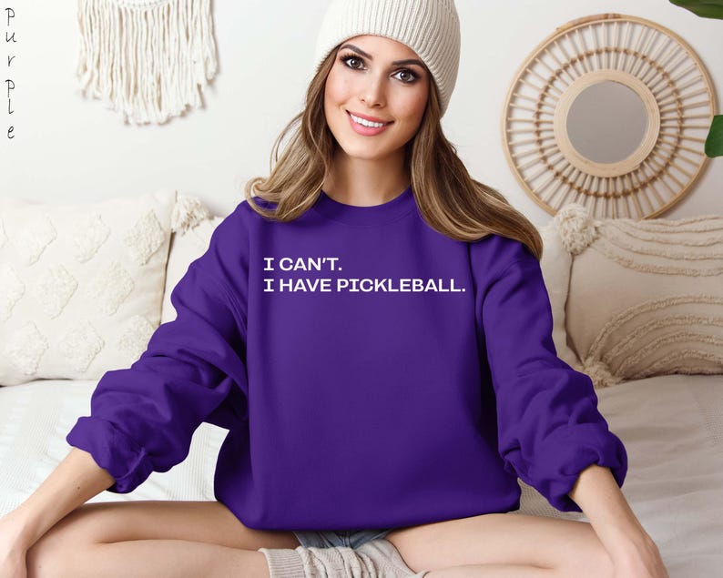 Pode incluir: Um moletom roxo escuro com o texto branco "I CAN'T. I HAVE PICKLEBALL.". A parte superior de manga comprida &eacute; de cor lisa. A imagem mostra o moletom.
