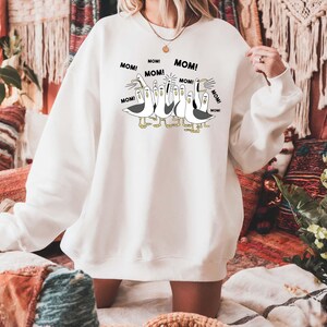May include: White sweatshirt featuring a cartoon illustration of seagulls with the word "MOM!" in speech bubbles. The seagulls are in black and white with yellow feet and beaks. The sweatshirt is a relaxed fit.
