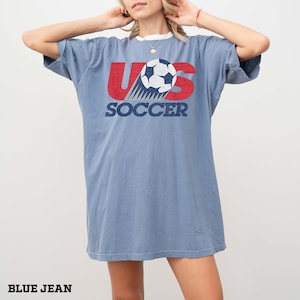 Pu&ograve; includere: Una t-shirt a maniche corte color blu jeans con una grafica sul petto. La grafica mostra il testo US in lettere rosse consumate con un pallone da calcio che sostituisce la lettera S. Sotto la grafica, la parola SOCCER &egrave; stampata in blu navy.