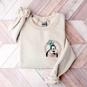 Op de afbeelding: Een cr&egrave;mekleurige sweatshirt met een cartoonafbeelding van Goofy. De afbeelding is een cirkelvormig ontwerp met een geblokte achtergrond. Goofy draagt een blauwe hoed en heeft een grote, open mond. De sweatshirt heeft een ronde hals en lange mouwen.