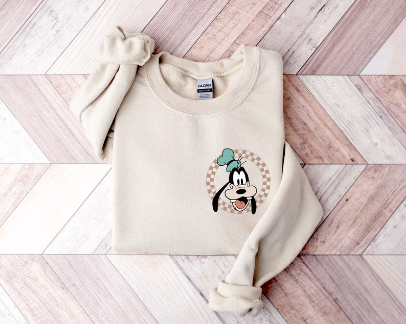 Op de afbeelding: Een cr&egrave;mekleurige sweatshirt met een cartoonafbeelding van Goofy. De afbeelding is een cirkelvormig ontwerp met een geblokte achtergrond. Goofy draagt een blauwe hoed en heeft een grote, open mond. De sweatshirt heeft een ronde hals en lange mouwen.