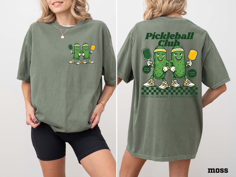 May include: A moss green t-shirt featuring a small graphic on the front left chest of two cartoon pickles holding pickleball paddles. The back displays a large graphic with the text Pickleball Club, two pickles, and the words Official Member Weekend League.