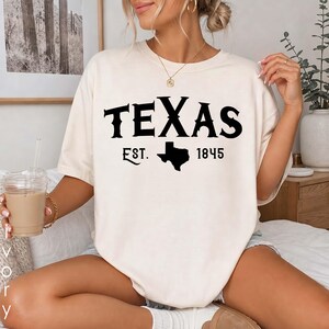 K&ouml;nnte beinhalten: Ein cremefarbenes Kurzarm-T-Shirt mit dem schwarzen Schriftzug TEXAS, der Abk&uuml;rzung EST., der Jahreszahl 1845 und einer Silhouette des Bundesstaates Texas. Das Shirt hat einen Rundhalsausschnitt und eine lockere, entspannte Passform.