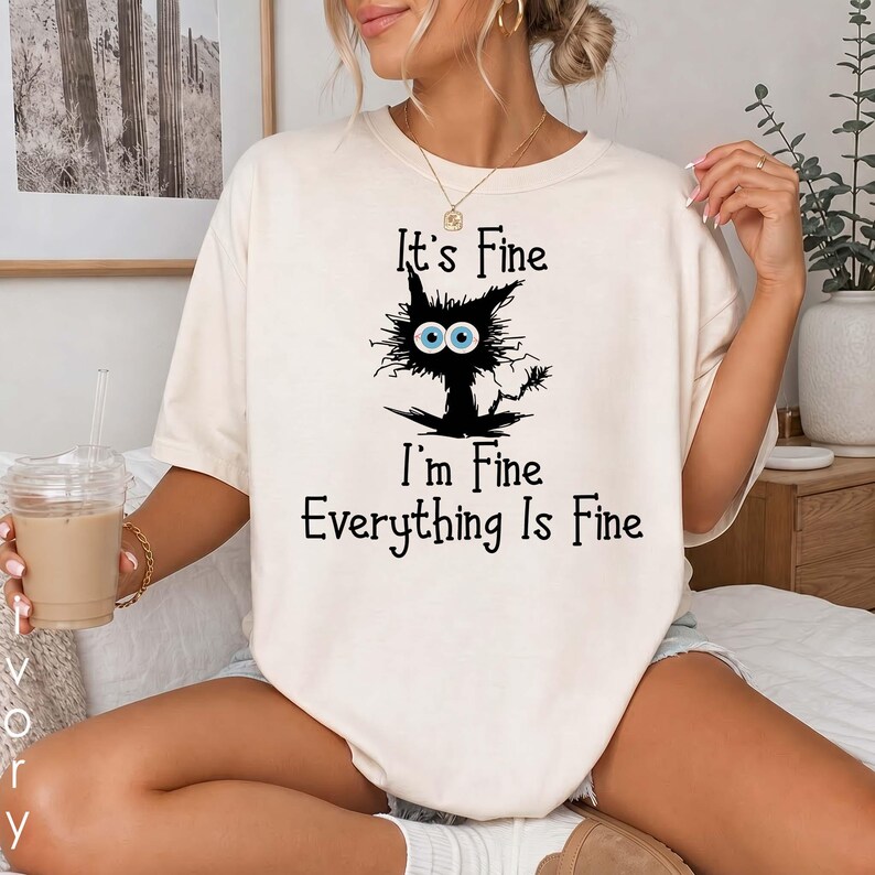 May include: A cream-colored short-sleeve t-shirt featuring a black graphic of a messy-haired cat with large blue eyes. The text printed in black reads: It's Fine, I'm Fine, Everything Is Fine. The shirt has a crew neckline and a relaxed fit.