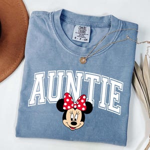May include: A blue Comfort Colors t-shirt with the word "AUNTIE" in white block letters and a Minnie Mouse graphic. The Minnie Mouse graphic has a red bow with white polka dots. A gold necklace is draped across the shirt.