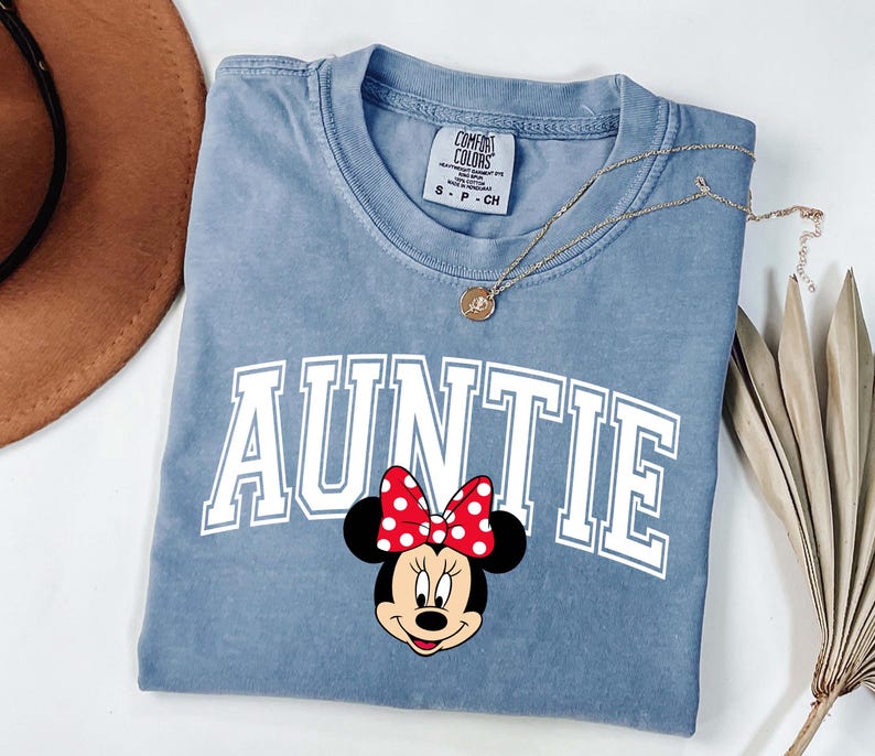 May include: A blue Comfort Colors t-shirt with the word "AUNTIE" in white block letters and a Minnie Mouse graphic. The Minnie Mouse graphic has a red bow with white polka dots. A gold necklace is draped across the shirt.