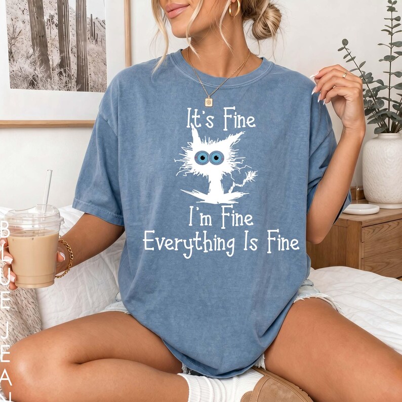 May include: A blue short-sleeve t-shirt featuring a white graphic of a wide-eyed, disheveled cat. The text printed in white reads, "It's Fine, I'm Fine, Everything Is Fine." The shirt has a crew neckline and a relaxed, loose-fitting silhouette.