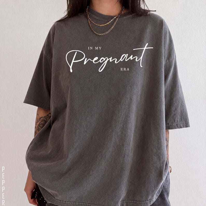 May include: A charcoal gray short-sleeve t-shirt featuring the white text "IN MY Pregnant ERA" printed across the chest. The shirt has a relaxed, oversized fit with a crew neckline and a washed, vintage-style fabric texture.