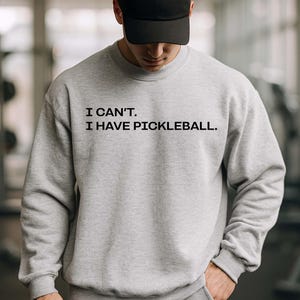 Pode incluir: Um conjunto de moletom cinza claro e cal&ccedil;as de moletom combinando. O moletom tem o texto "I CAN'T. I HAVE PICKLEBALL." em preto. Um bon&eacute; de beisebol preto completa o traje. A pessoa est&aacute; em uma sala bem iluminada.
