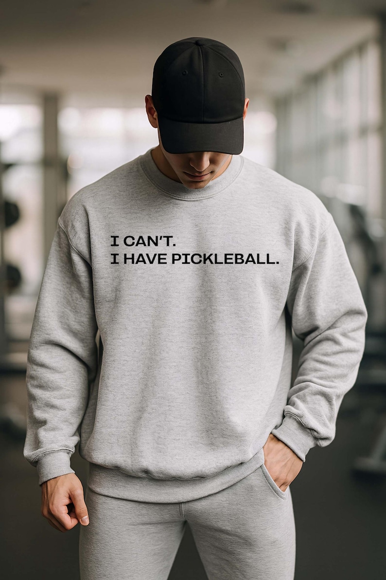 Pode incluir: Um conjunto de moletom cinza claro e cal&ccedil;as de moletom combinando. O moletom tem o texto "I CAN'T. I HAVE PICKLEBALL." em preto. Um bon&eacute; de beisebol preto completa o traje. A pessoa est&aacute; em uma sala bem iluminada.