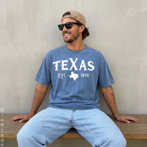 K&ouml;nnte beinhalten: Ein blaues kurz&auml;rmeliges T-Shirt mit dem wei&szlig;en Schriftzug TEXAS, der Abk&uuml;rzung EST., der Zahl 1845 und einer wei&szlig;en Silhouette des Bundesstaates Texas. Das T-Shirt wird mit einer hellblauen Jeans, einer beigen Baseballkappe und einer schwarzen Sonnenbrille getragen.