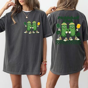 May include: A charcoal gray t-shirt featuring a graphic of two cartoon pickles wearing headbands and sneakers, each holding a pickleball paddle. The back of the shirt displays the same graphic with the text "Pickleball Club" in dark green letters.
