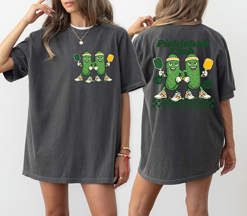 May include: A charcoal gray t-shirt featuring a graphic of two cartoon pickles wearing headbands and sneakers, each holding a pickleball paddle. The back of the shirt displays the same graphic with the text "Pickleball Club" in dark green letters.