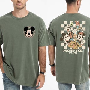 Op de afbeelding: Mosgroen t-shirt met Mickey Mouse op de voorkant en een groep Disney-personages op de achterkant. Het shirt heeft een klassieke pasvorm en is gemaakt van een zacht, comfortabel materiaal. De achterkant van het shirt heeft Mickey & Co. Est. 1928.