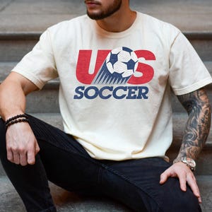 Pu&ograve; includere: Una t-shirt a maniche corte color avorio con un design grafico che presenta le lettere rosse US, un pallone da calcio bianco e nero con linee di movimento e la parola SOCCER in lettere maiuscole blu al centro del petto.