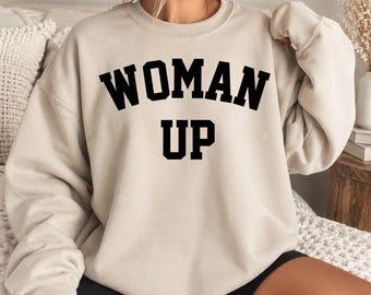 May include: A sand-colored crewneck sweatshirt featuring the text "WOMAN UP" in bold, black, collegiate-style lettering across the chest. The garment has long sleeves, a ribbed neckline, and a relaxed fit, providing a simple and clean look.