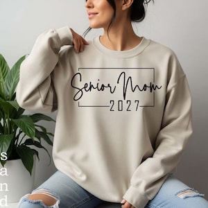 Może przedstawiać: Beżowa bluza z okrągłym dekoltem, ozdobiona czarną prostokątną ramką z napisem Senior Mom oraz rokiem 2027, wykonanymi mieszanką czcionek kursywnych i bezszeryfowych. Odzież posiada długie rękawy oraz ściągacze przy dekolcie, mankietach i u dołu.