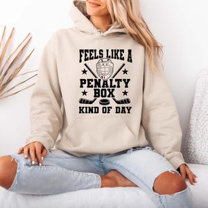 A woman wearing a beige hoodie with a graphic design that says "FEELS LIKE A PENALTY BOX KIND OF DAY" is sitting on a couch.