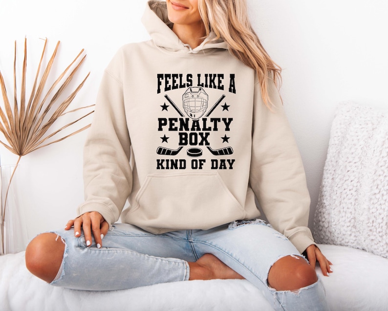 A woman wearing a beige hoodie with a graphic design that says "FEELS LIKE A PENALTY BOX KIND OF DAY" is sitting on a couch.