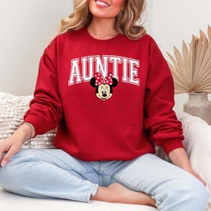 May include: A red sweatshirt with the word "AUNTIE" in white block letters. A Minnie Mouse graphic with a red bow is centered below the word. The sweatshirt is long-sleeved and made of a soft material.