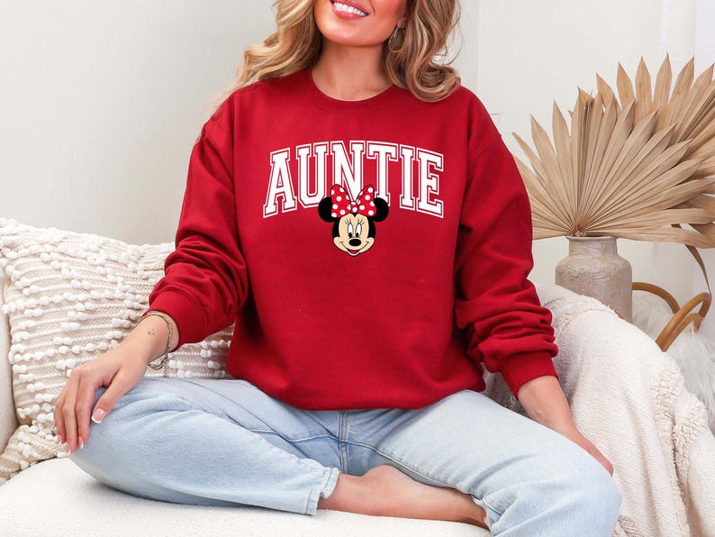 May include: A red sweatshirt with the word "AUNTIE" in white block letters. A Minnie Mouse graphic with a red bow is centered below the word. The sweatshirt is long-sleeved and made of a soft material.