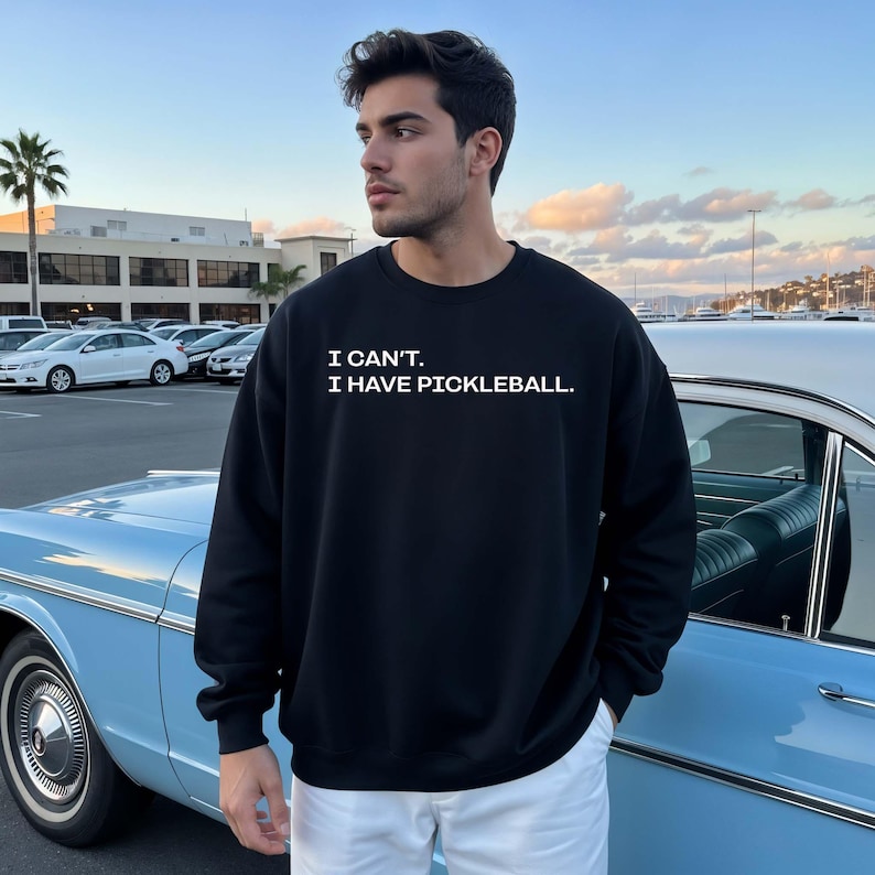 Pode incluir: Camisola preta com o texto branco "I CAN'T. I HAVE PICKLEBALL.". Uma pessoa usa a camisola perto de um carro cl&aacute;ssico azul claro. O fundo mostra um estacionamento e edif&iacute;cios.