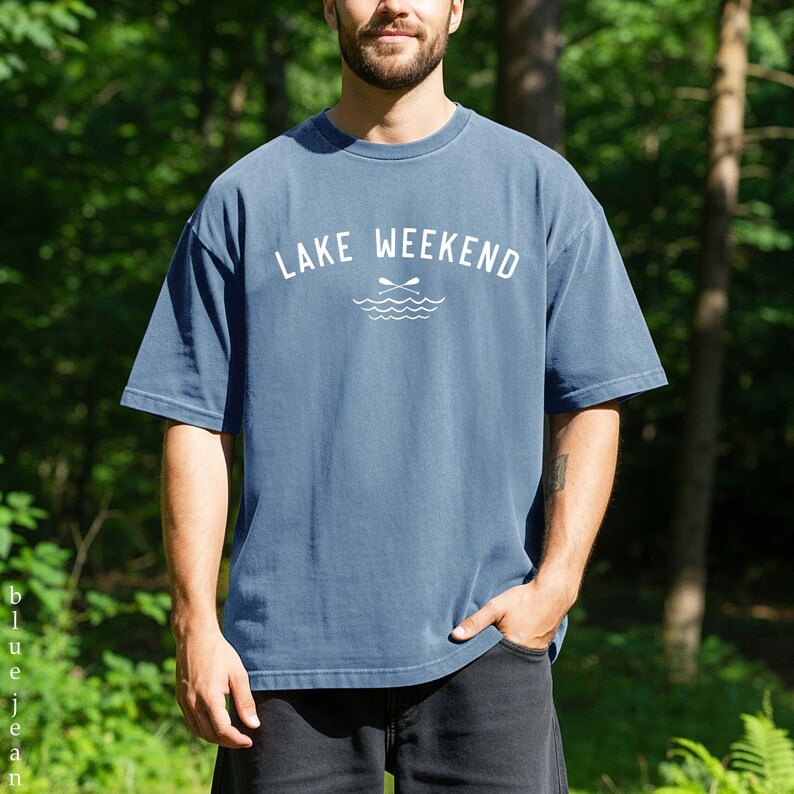 May include: A slate blue short-sleeve t-shirt featuring the white text "LAKE WEEKEND" printed across the chest, accompanied by a graphic of two crossed oars positioned above stylized wavy lines representing water.