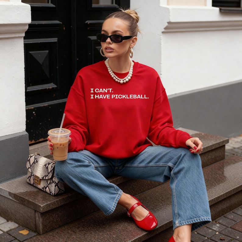 Pode incluir: Uma camisola vermelha com o texto "I CAN'T. I HAVE PICKLEBALL." em branco. A pessoa est&aacute; usando jeans azuis e sapatos vermelhos. Um colar de p&eacute;rolas, &oacute;culos de sol e uma bolsa de pele de cobra completam o look. Uma x&iacute;cara transparente de caf&eacute; est&aacute; na m&atilde;o da pessoa.