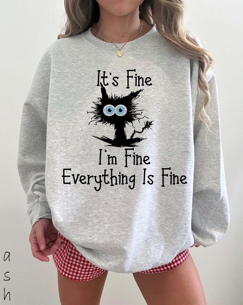 May include: A light gray crewneck sweatshirt featuring a black graphic of a messy-haired cat with large blue eyes. The text "It's Fine, I'm Fine, Everything Is Fine" is printed in black lettering above and below the cat illustration.