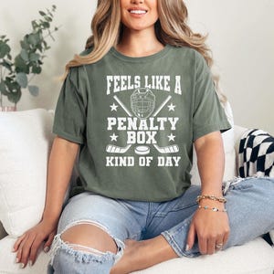 May include: A moss green t-shirt with the text "FEELS LIKE A PENALTY BOX KIND OF DAY" in white. The graphic includes a hockey helmet, crossed hockey sticks, and a hockey puck. The shirt is worn with blue jeans.