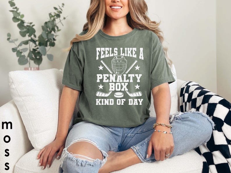 May include: A moss green t-shirt with the text "FEELS LIKE A PENALTY BOX KIND OF DAY" in white. The graphic includes a hockey helmet, crossed hockey sticks, and a hockey puck. The shirt is worn with blue jeans.