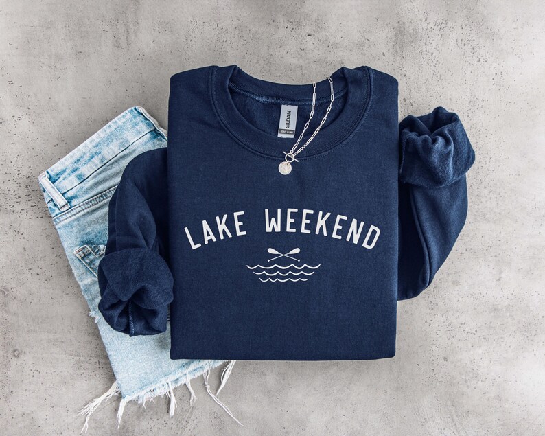 May include: A navy blue crewneck sweatshirt featuring the white text LAKE WEEKEND arched above a graphic of two crossed oars and stylized water waves. A silver chain necklace rests on the collar, and light blue denim jeans are visible underneath.