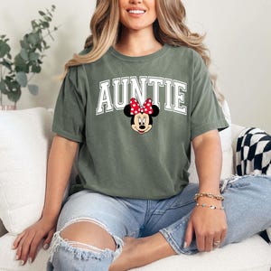May include: Olive green t-shirt with the word "AUNTIE" in white block letters and a Minnie Mouse graphic. The shirt is made of a soft material and is a casual style.