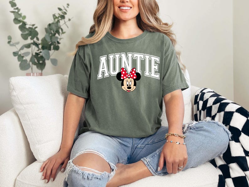 May include: Olive green t-shirt with the word "AUNTIE" in white block letters and a Minnie Mouse graphic. The shirt is made of a soft material and is a casual style.